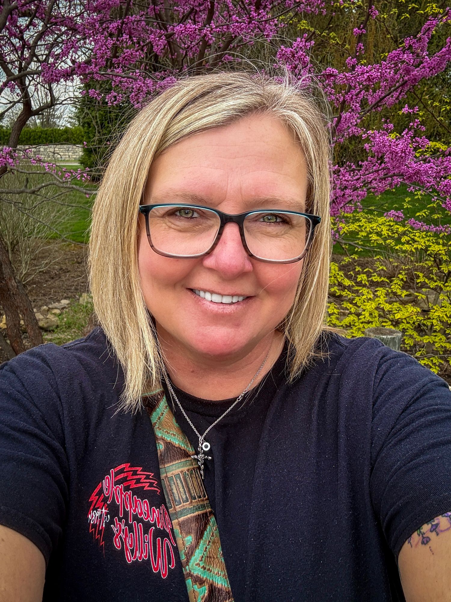Smiling blonde woman with glasses taking a selfie outdoors with purple redbud trees in spring bloom behind her.