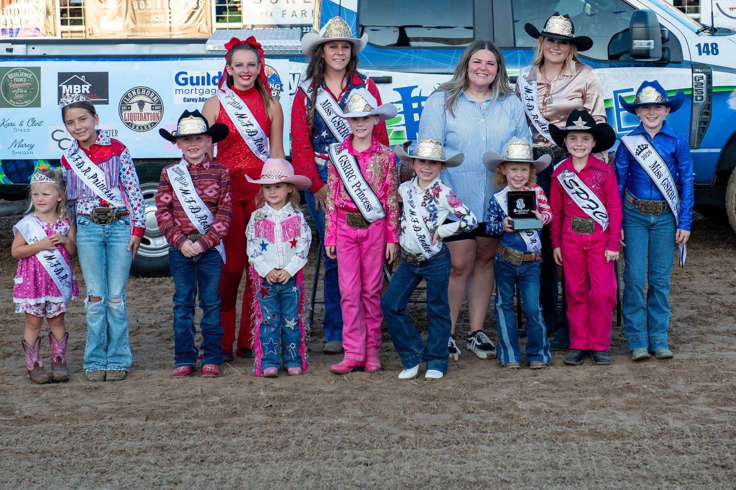 Group photo of rodeo royalty queens and young contestants posing together at an outdoor rodeo event.