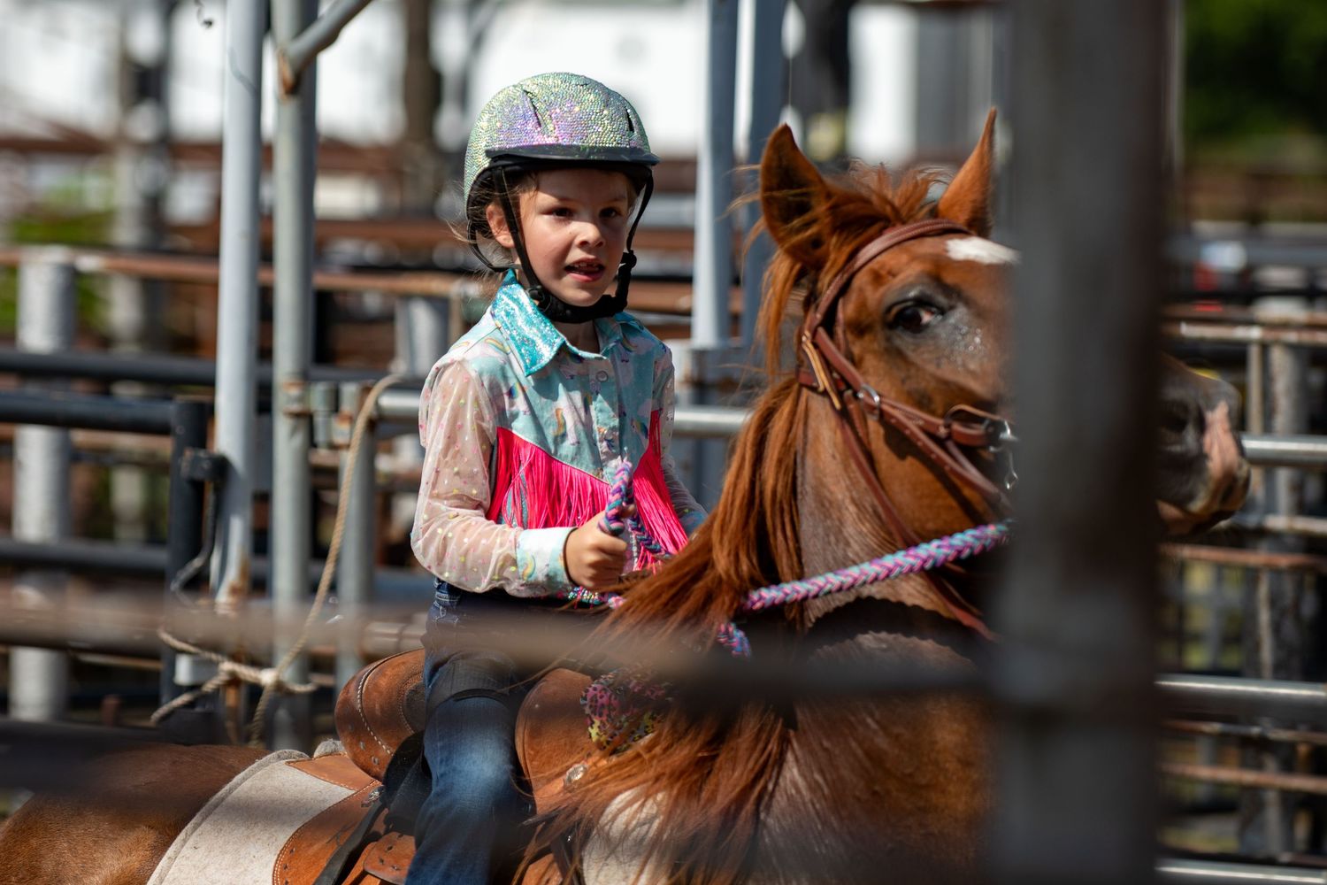 Young child wearing helmet riding a chestnut horse in a rodeo arena behind a fence.