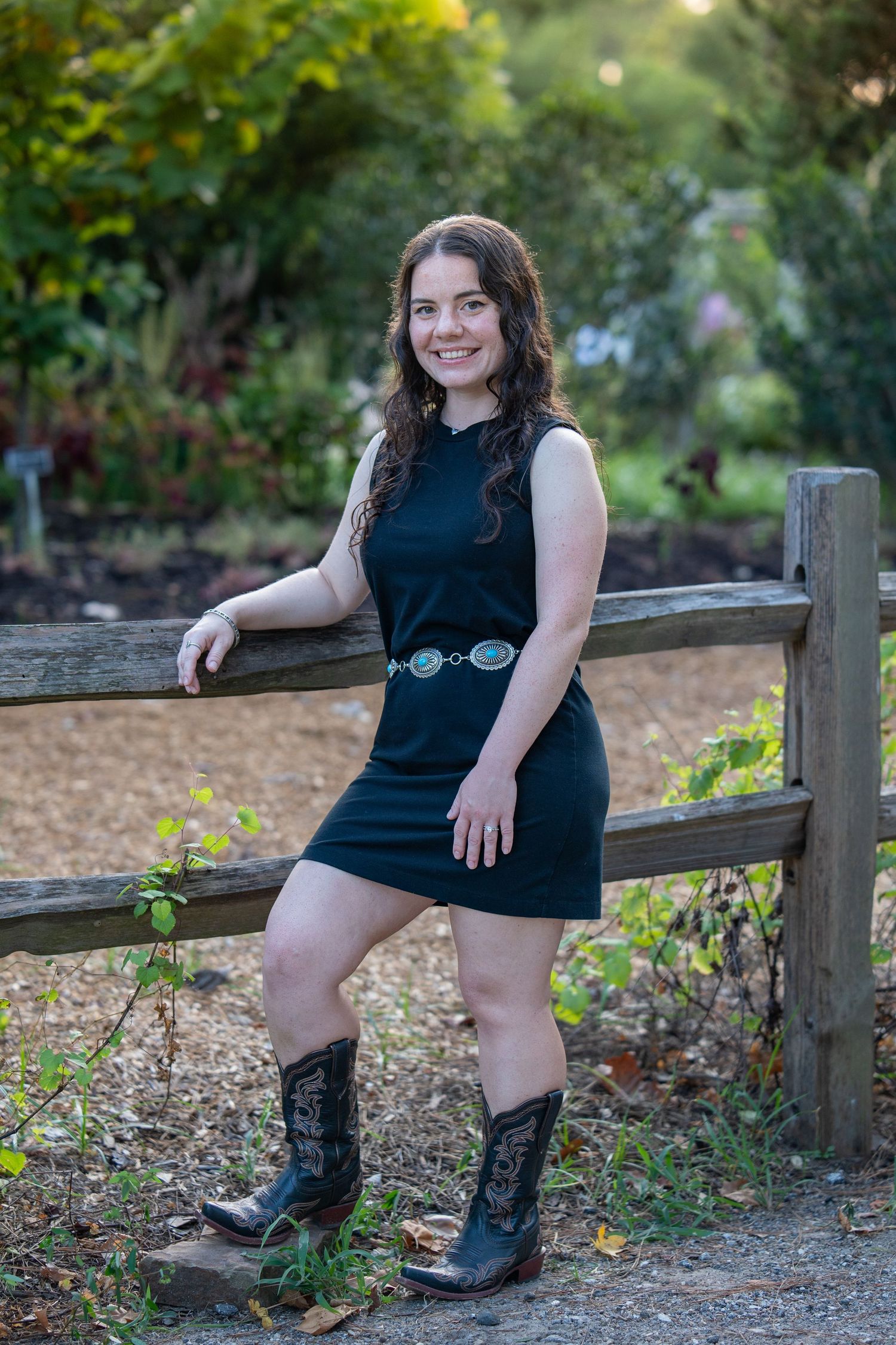 Young woman in black mini dress, decorative belt, and cowboy boots leaning on wooden fence in lush garden setting.