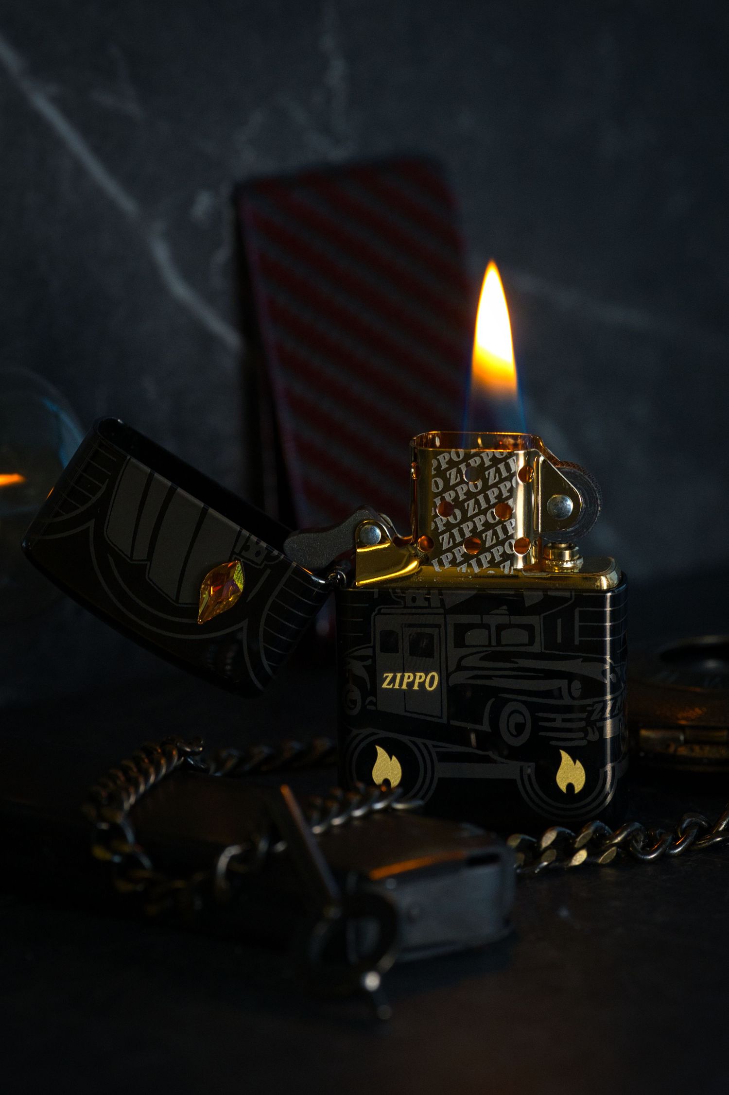 Zippo lighter with gold flame burning in dark moody setting with chain and red striped box.