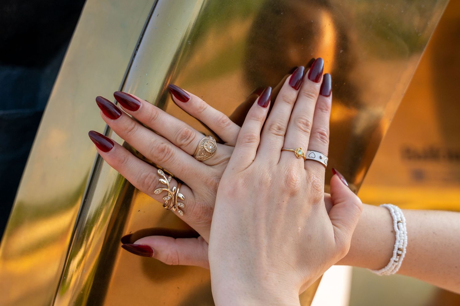 Close-up of hands with dark red nails and gold rings pressed against a gold reflective surface.
