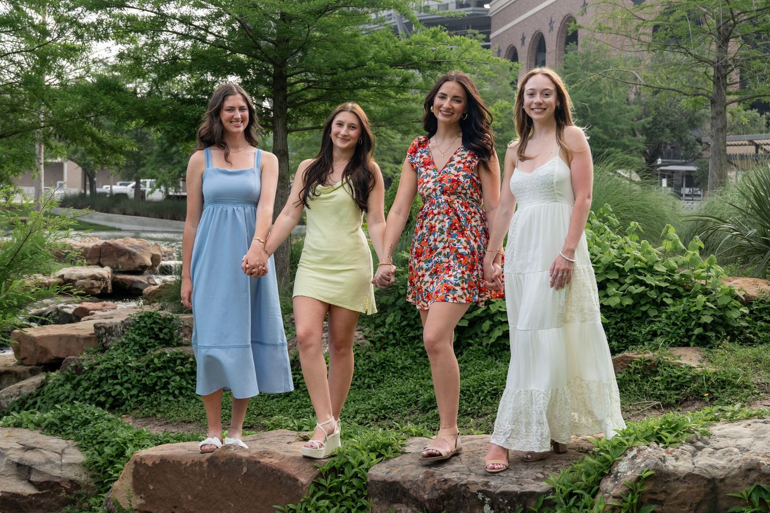 Four young women in colorful summer dresses holding hands and walking on rocks in a lush green garden.