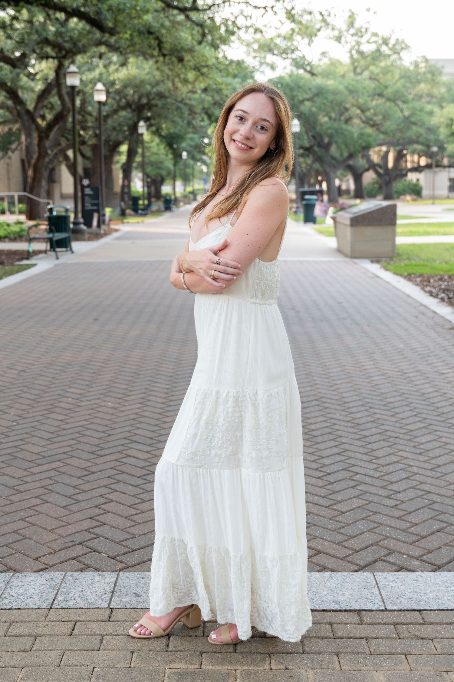 Young woman in a white lace maxi dress stands on a brick walkway lined with oak trees.