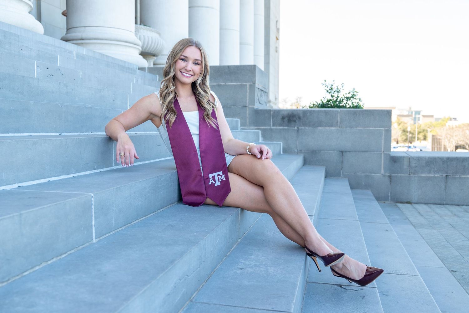 Smiling blonde woman in white outfit with Texas A&M maroon graduation stole on stone steps.