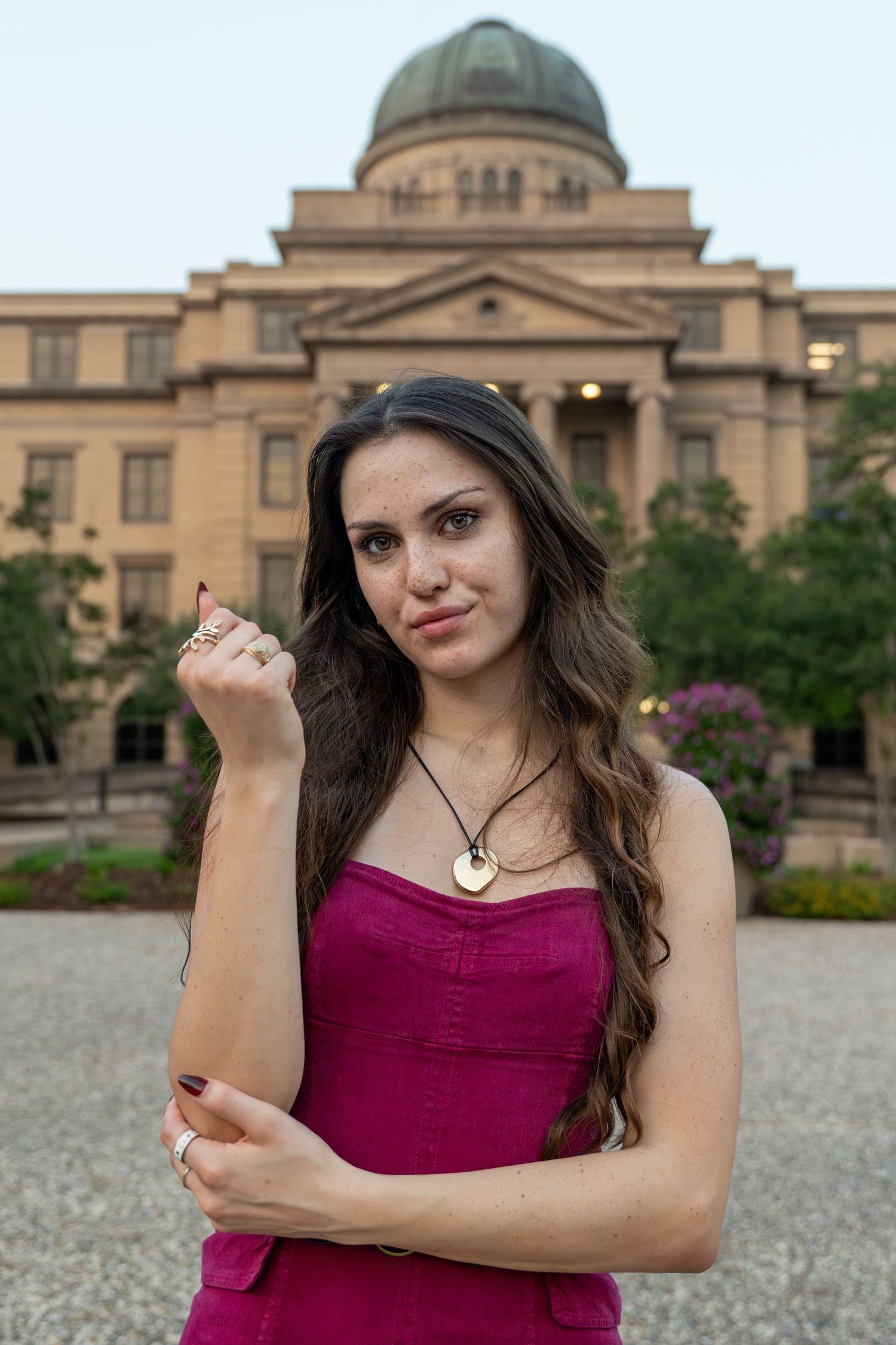 Young brunette woman in pink strapless dress posing in front of a domed university building at dusk.