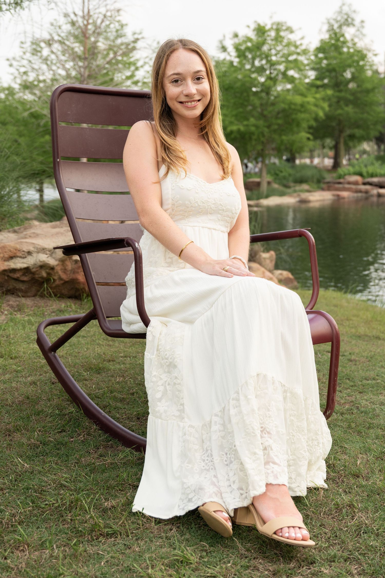 Young woman in a white lace maxi dress sits in a rocking chair beside a serene garden pond.