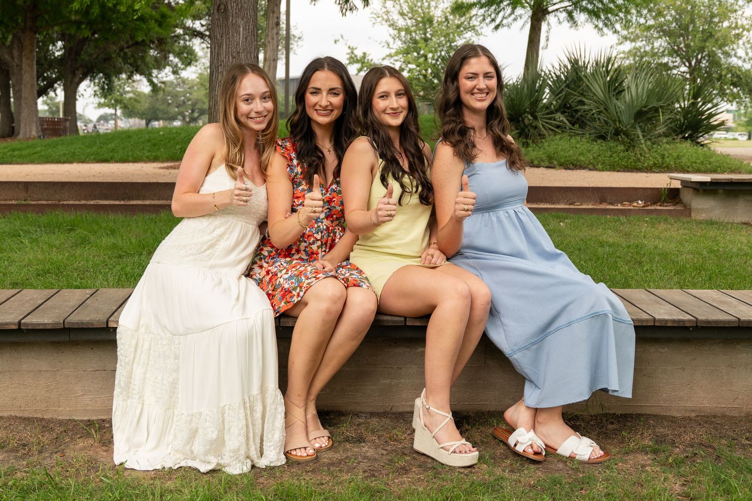 Four smiling young women in colorful summer dresses posing together on a park bench giving thumbs up.