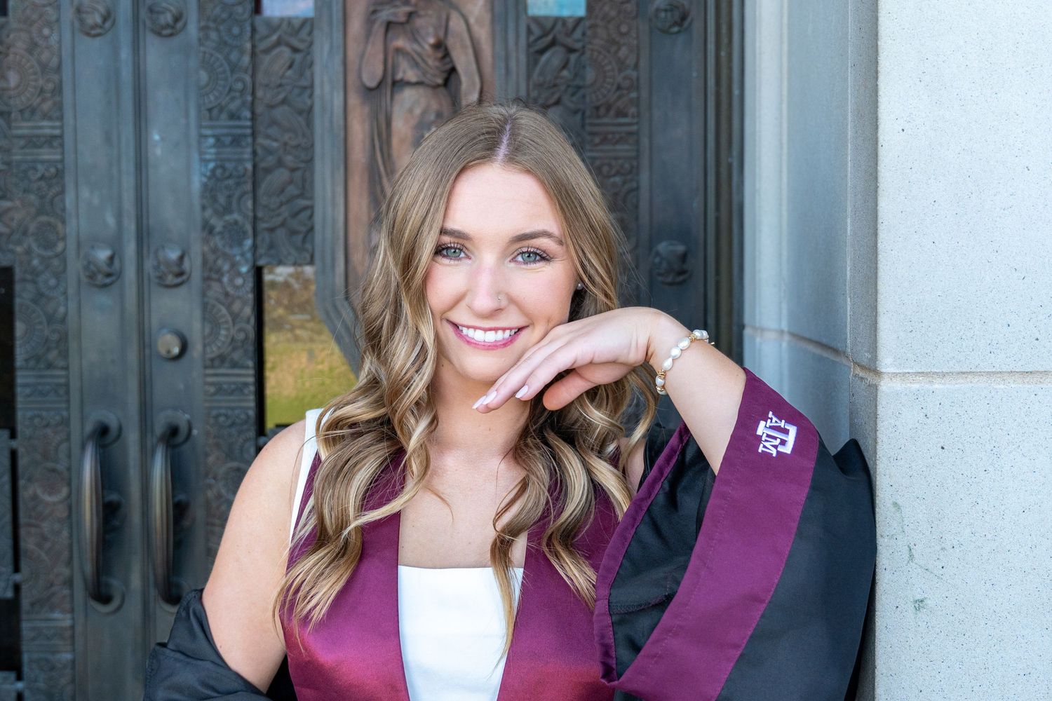 Smiling graduate wearing maroon sash and white top posing in front of ornate doors.