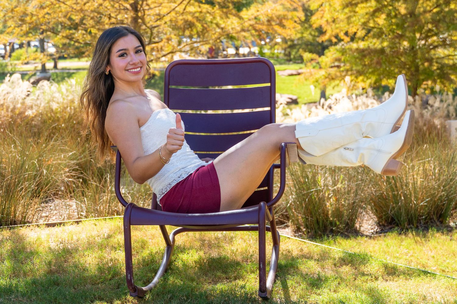 Young woman in white top, maroon shorts, and white boots relaxing in a purple chair outdoors in autumn.