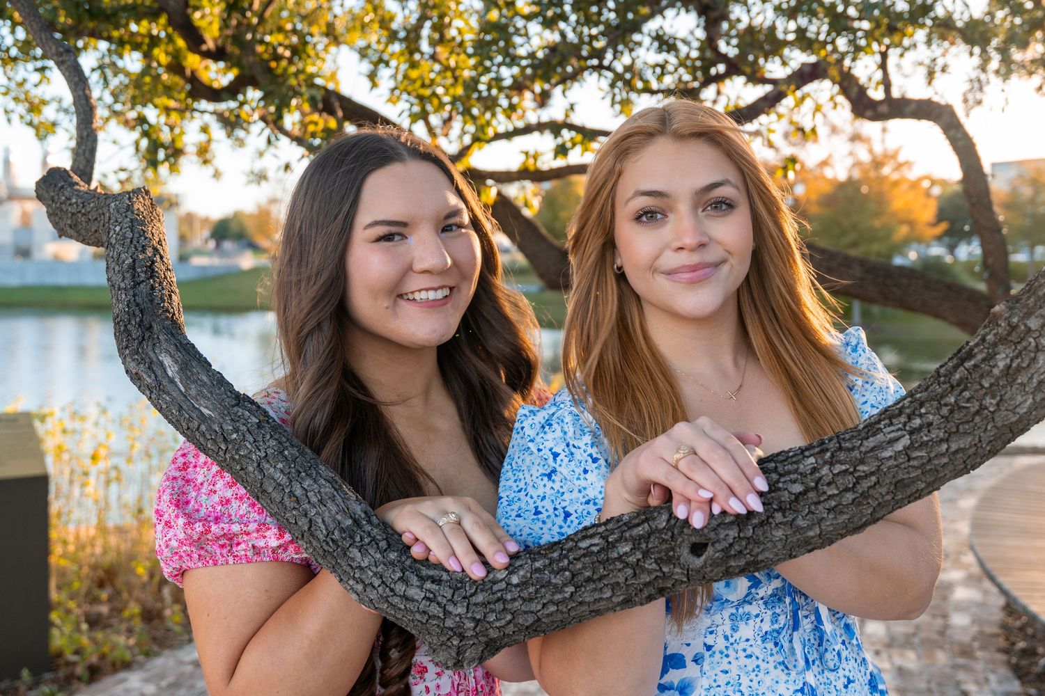 Two young women in floral dresses smiling and posing together leaning on a tree branch near a lake.