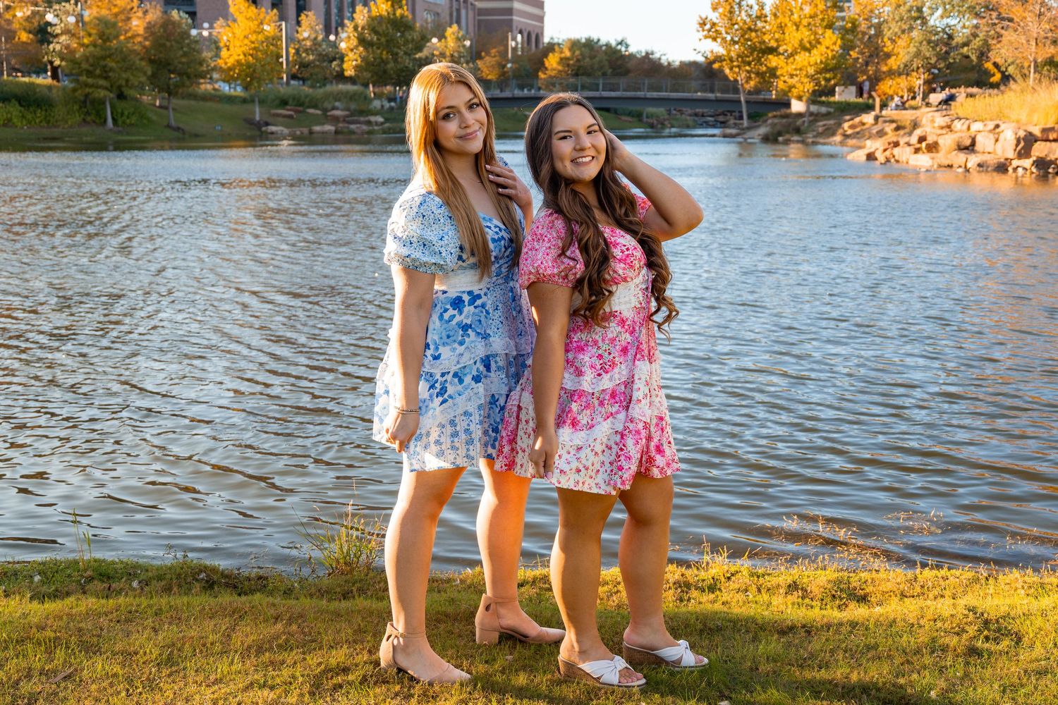 Two smiling women in colorful floral dresses stand together near a peaceful lakeside park setting.