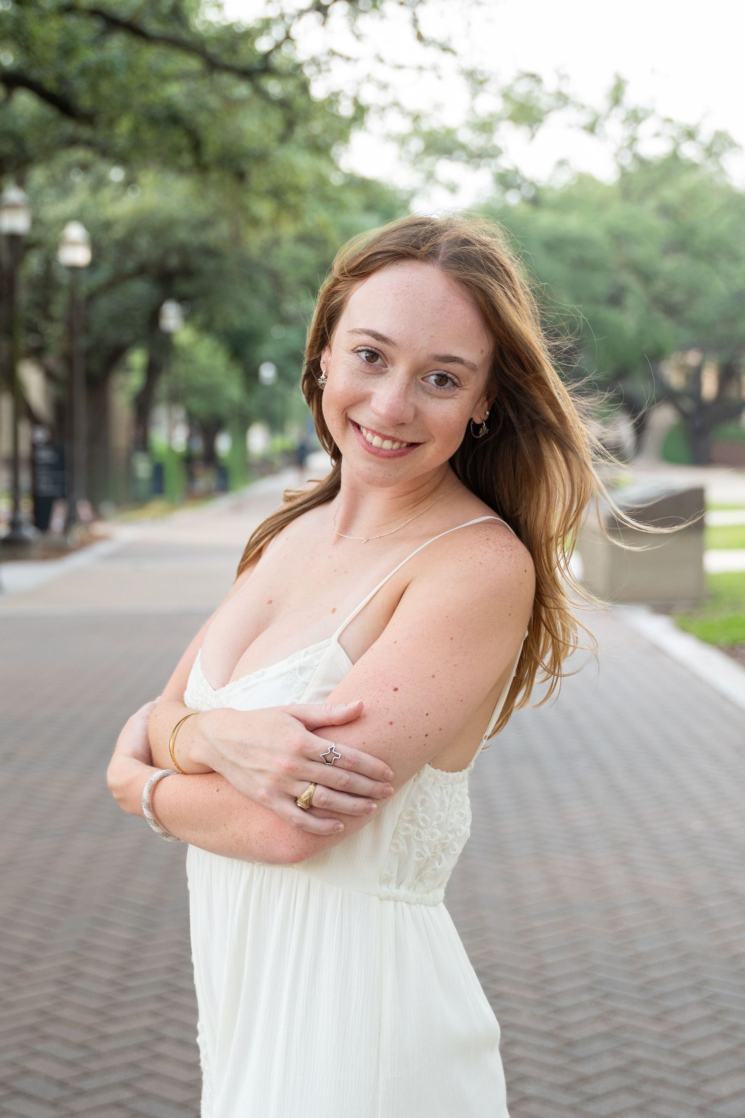 Smiling young woman in a white dress with arms crossed poses on a tree-lined brick pathway.