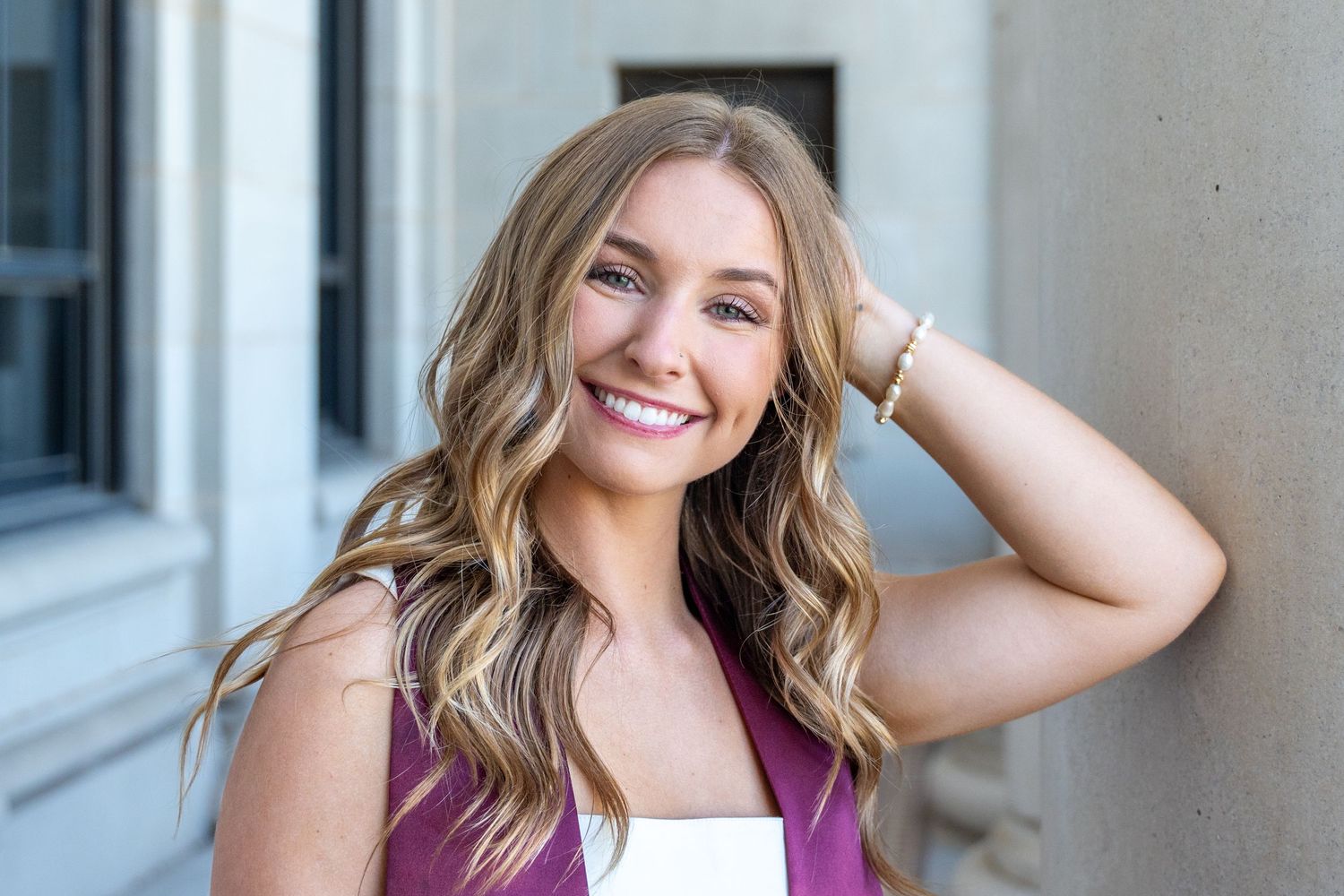 Smiling young woman with highlighted wavy hair in maroon top leaning against a column outdoors.
