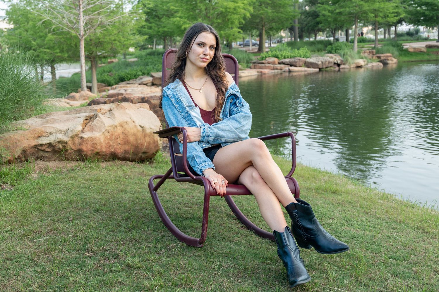 Woman in denim jacket and black boots sitting in rocking chair by a scenic lake.