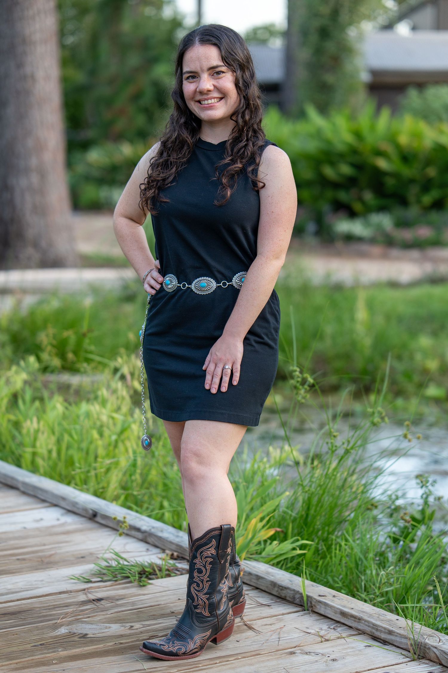 Woman in black sleeveless dress, turquoise belt, and cowboy boots smiling on a wooden walkway outdoors.