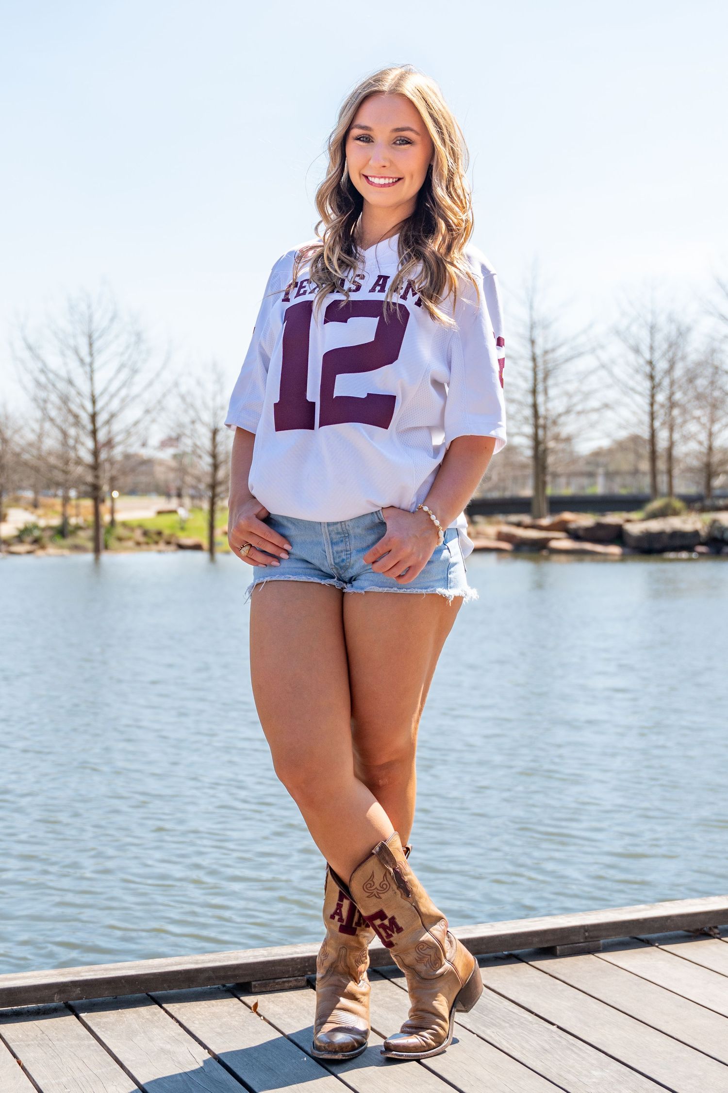 Smiling woman in Texas A&M number 12 jersey and cowboy boots stands on a dock by a lake.