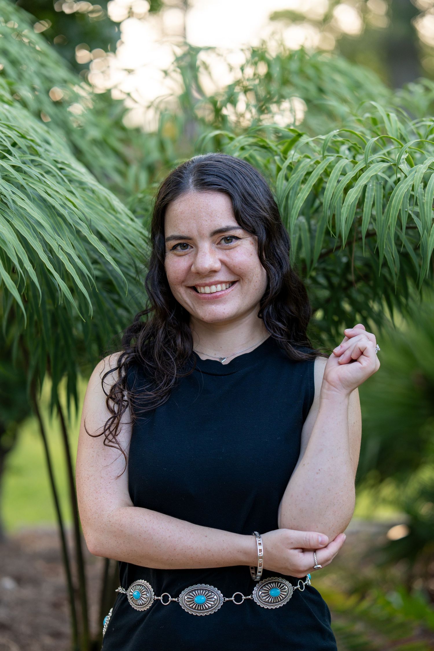 Smiling young woman in black sleeveless dress with turquoise belt, posing outdoors among lush green ferns.