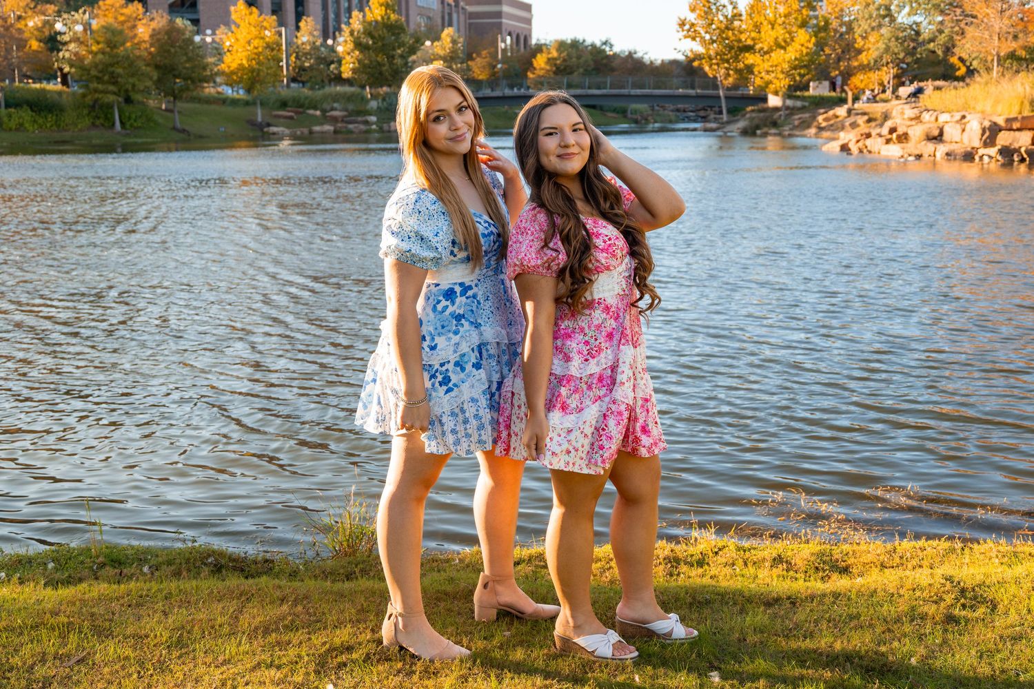 Two young women in floral dresses pose together by a scenic lake with fall foliage in the background.