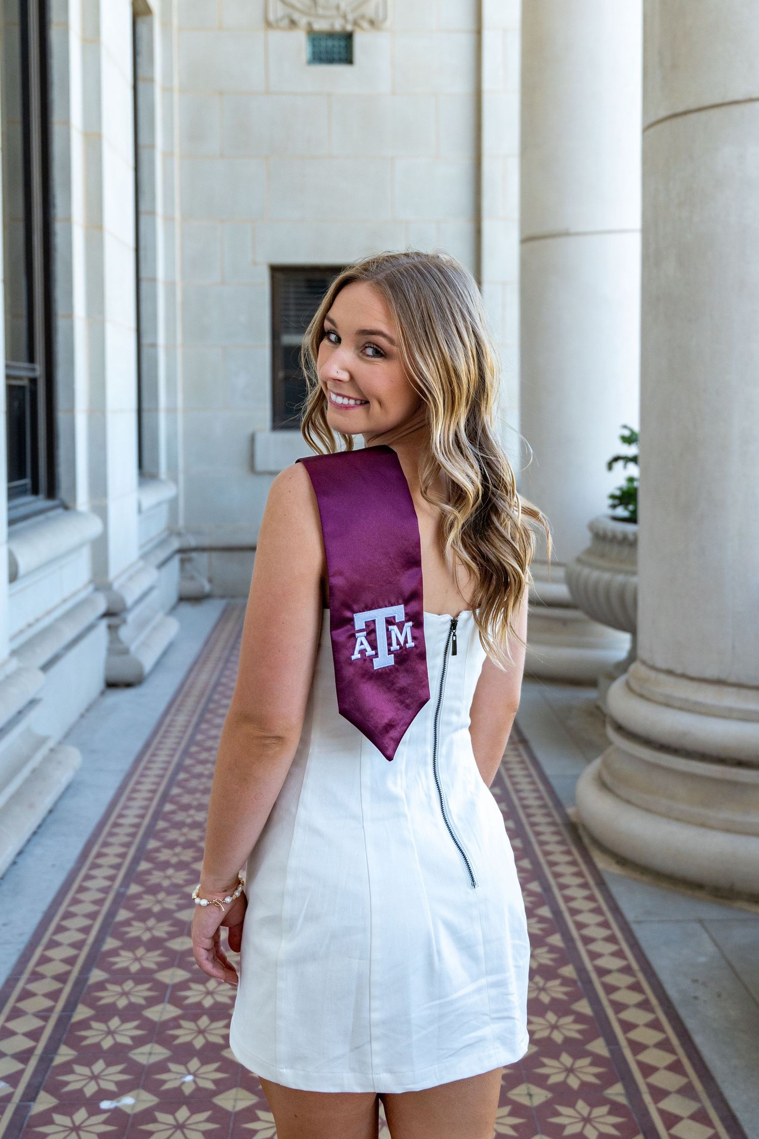 Smiling blonde woman in white dress wearing Texas A&M maroon graduation sash between columns.