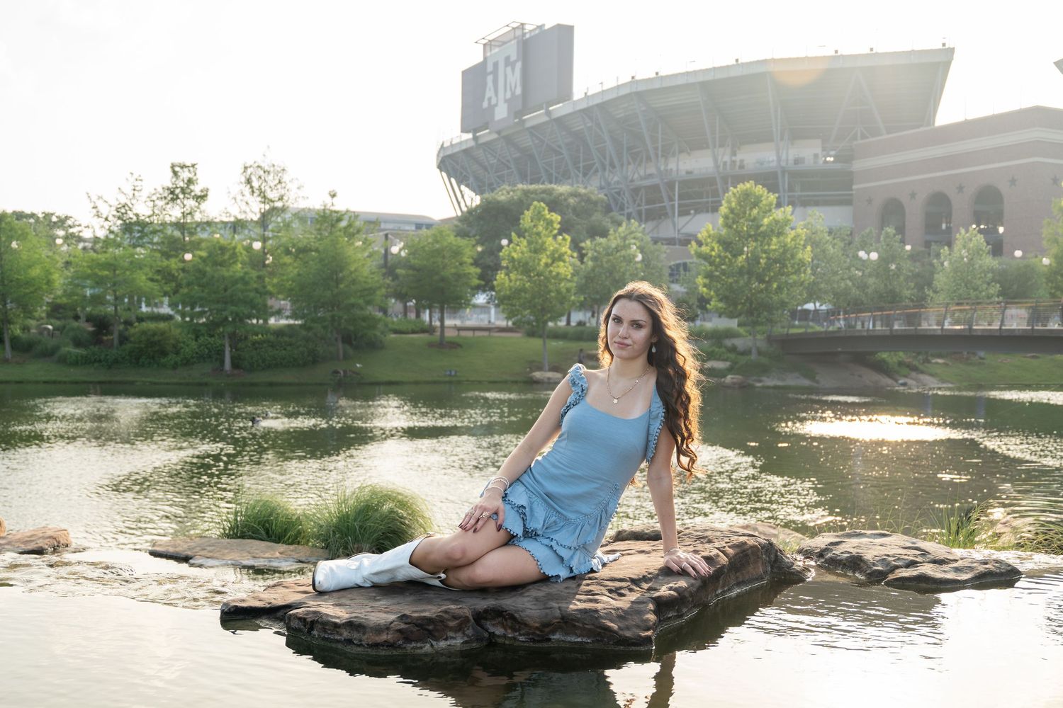 Young woman in blue dress and white boots sitting on a rock near a pond with a stadium backdrop.