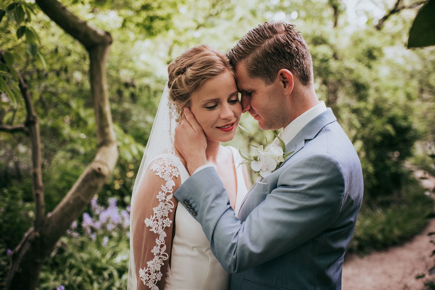 Bride and groom share tender forehead touch moment in lush green garden setting.
