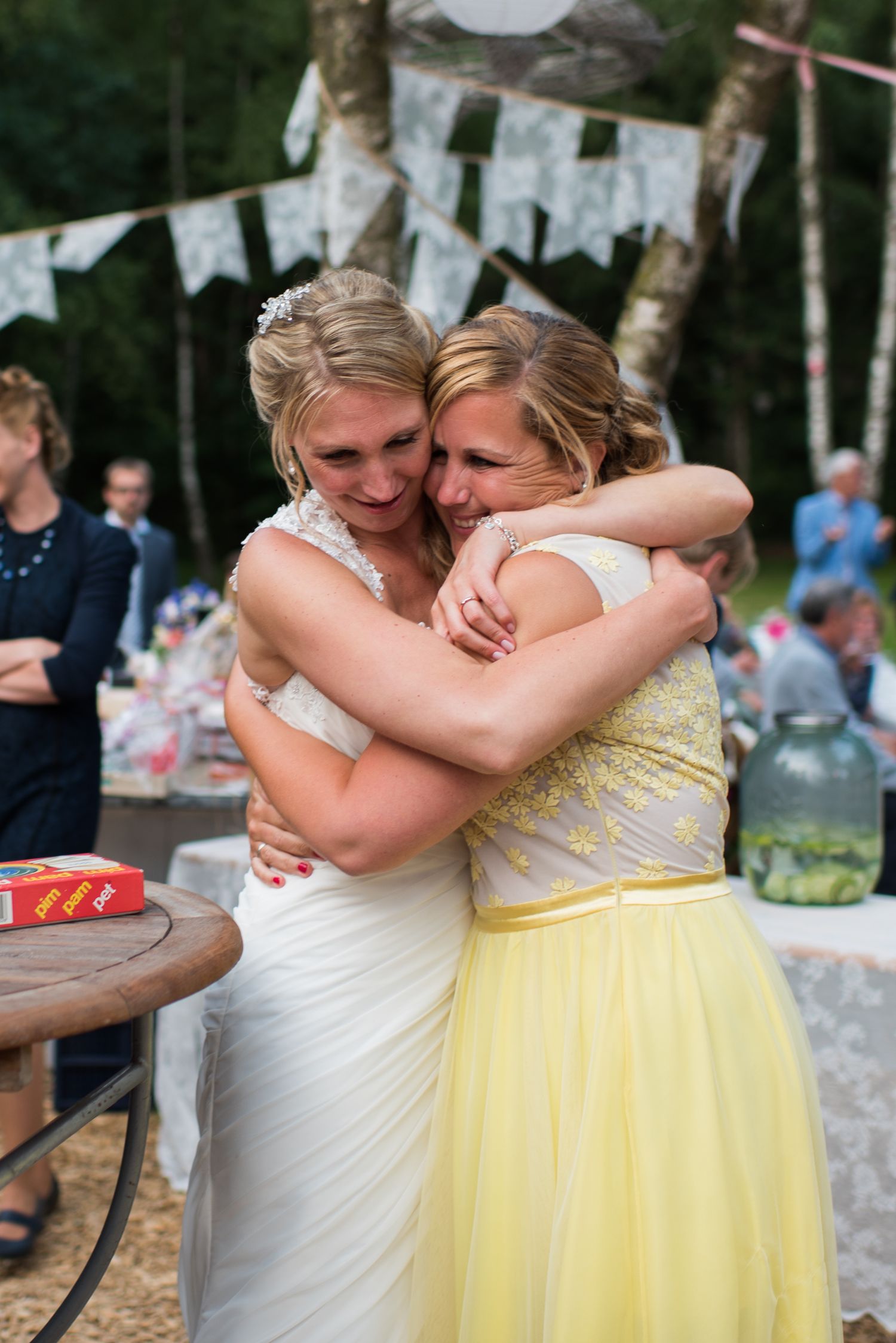 Bride in white gown shares an emotional embrace with bridesmaid in yellow dress at outdoor reception.