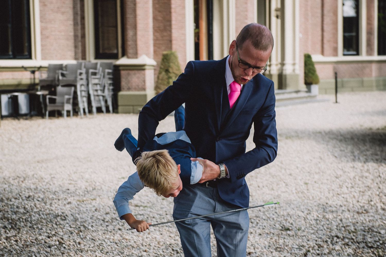 A man in a navy suit playfully lifts a young boy upside down on a gravel courtyard at a wedding. wedding photography Door Kim van de Broek