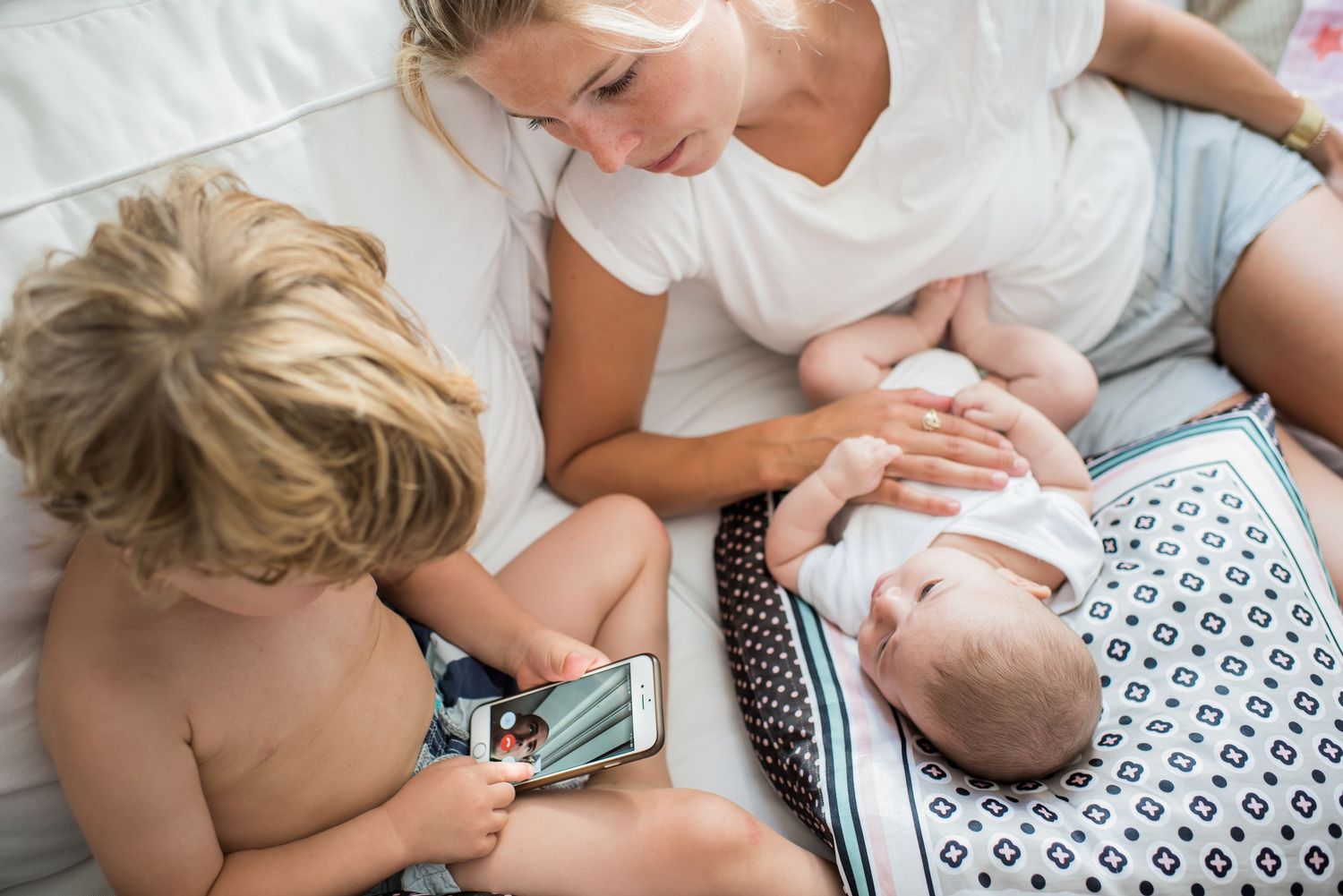 Mother sitting with toddler and newborn baby on a white bed at home.
