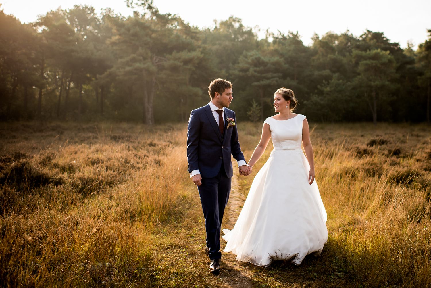 A bride and groom walk hand in hand through a golden field at sunset on their wedding day.