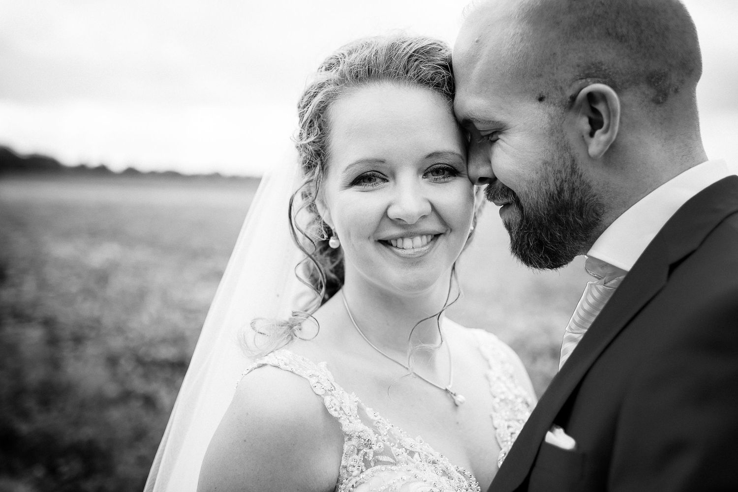 Close-up black and white portrait of smiling bride in lace gown with groom nuzzling her cheek outdoors.