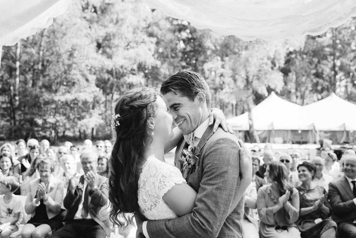 Black and white photo of bride and groom sharing first kiss at outdoor wedding ceremony with applauding guests.