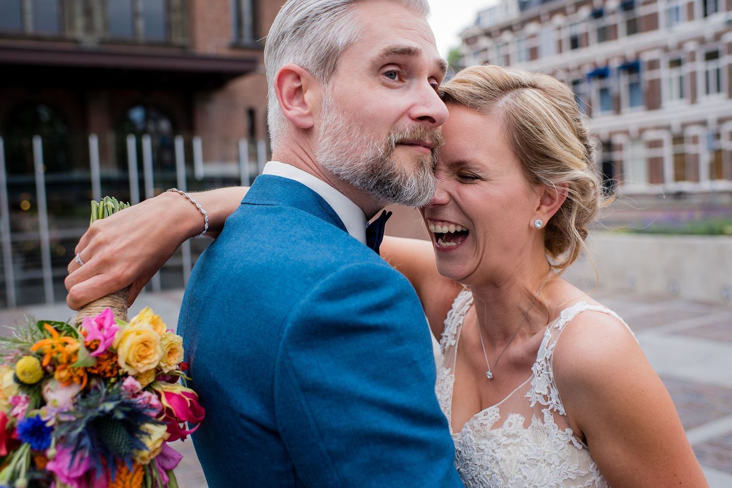Happy bride laughing as groom in blue suit embraces hergefotografeerd door documentaire trouwfotograaf Kim van den Broek