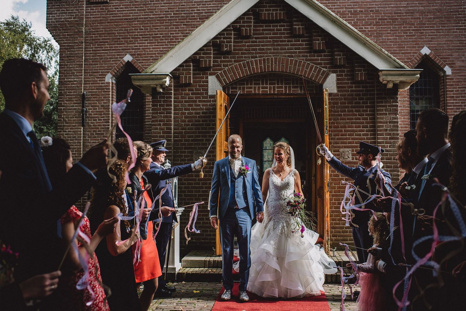 Newlyweds exit brick church as guests celebrate with colorful streamers on a sunny day. Door Kim van de Broek Fotografie