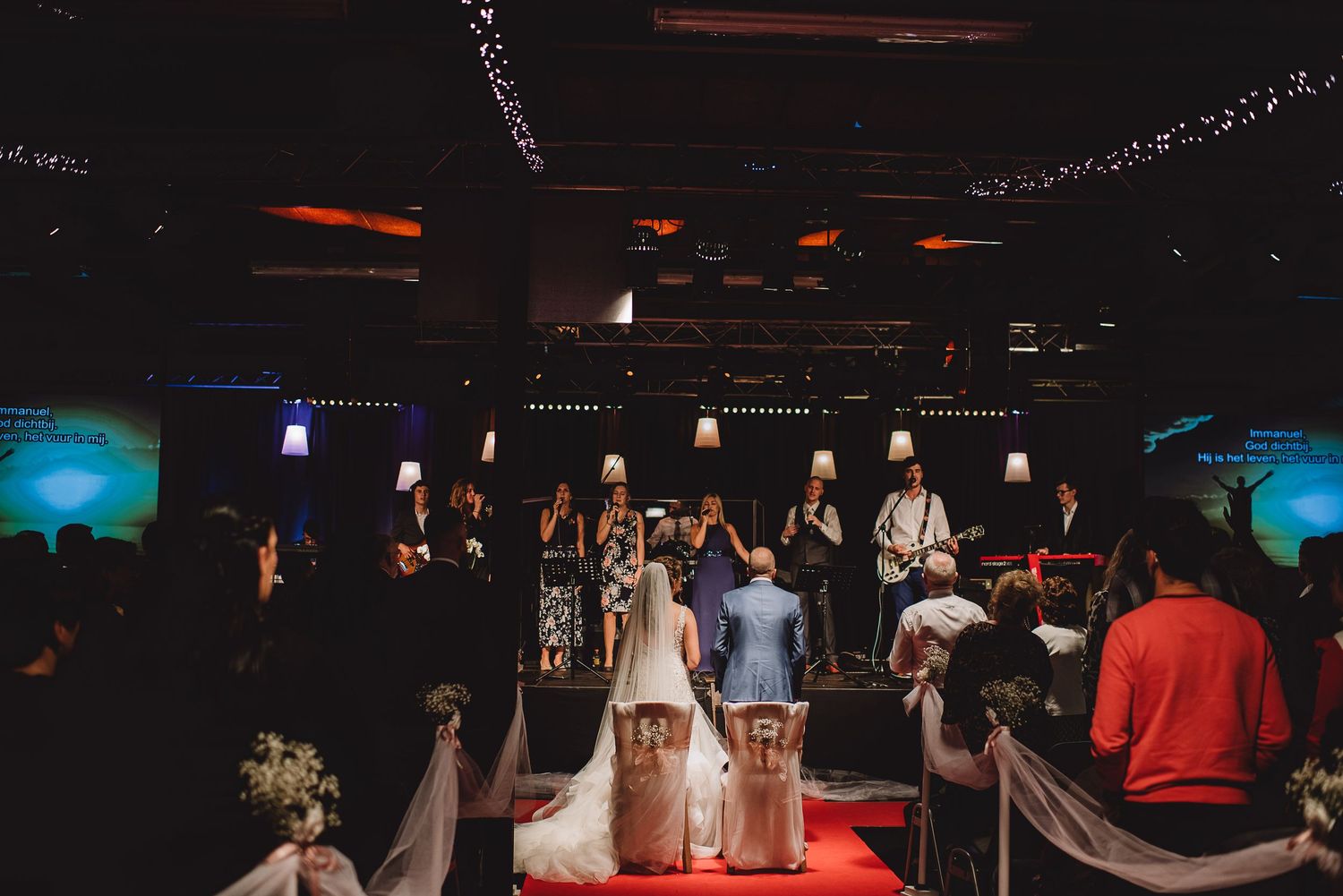 Wide shot of bride and groom kneeling before a live band during a church wedding ceremony. Door Kim van de Broek Fotografie
