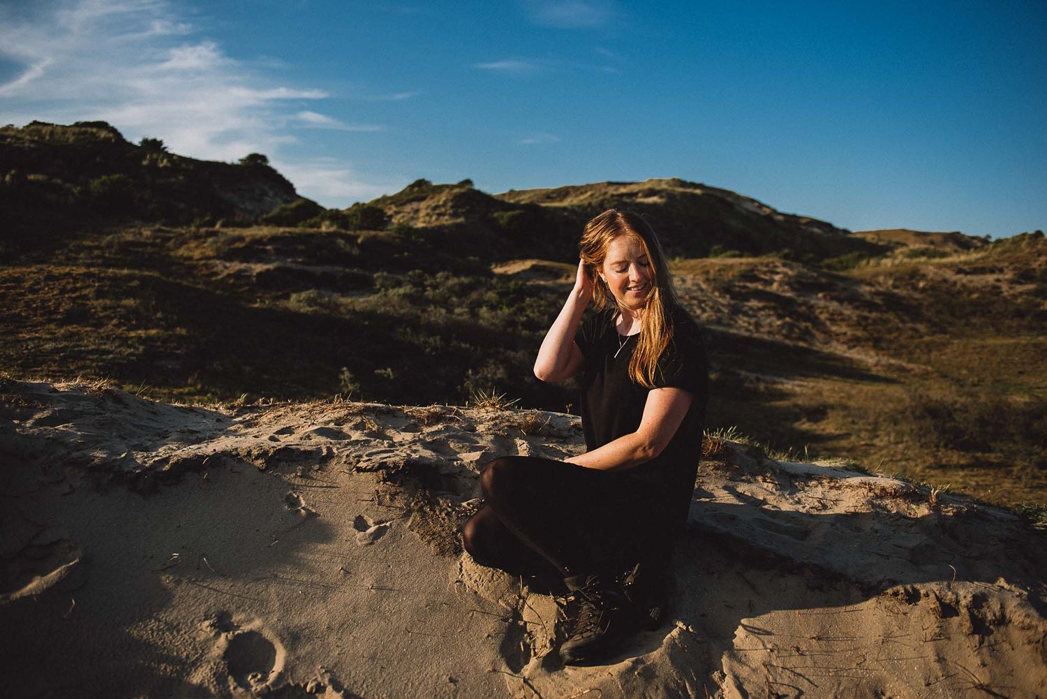Young blonde woman sitting on sandy desert rocks with dramatic hills and blue sky in the background.