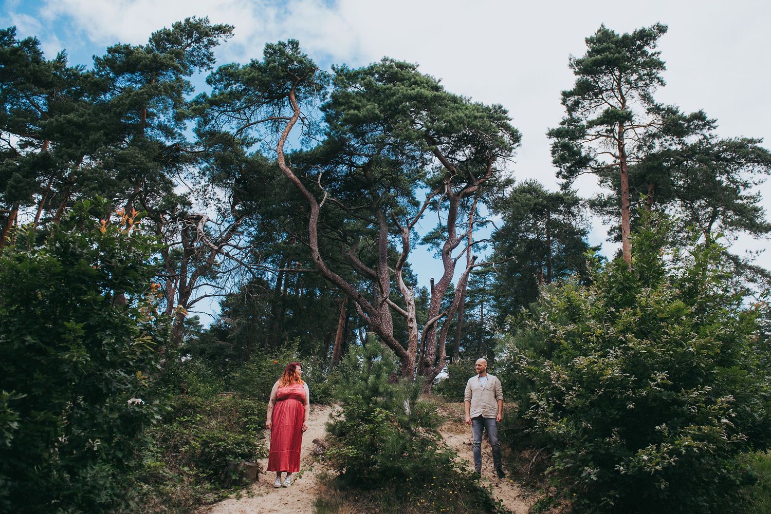 Couple standing apart in a lush forest with tall pine trees on a sandy path.