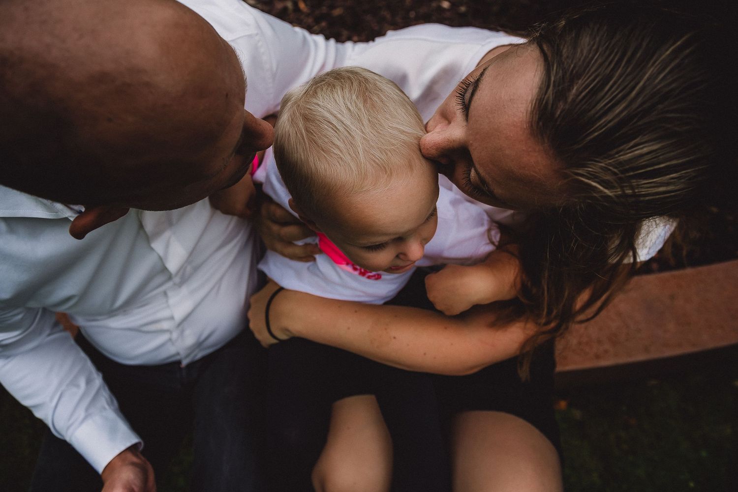 Aerial view of two parents leaning in to kiss their smiling toddler child from above.
