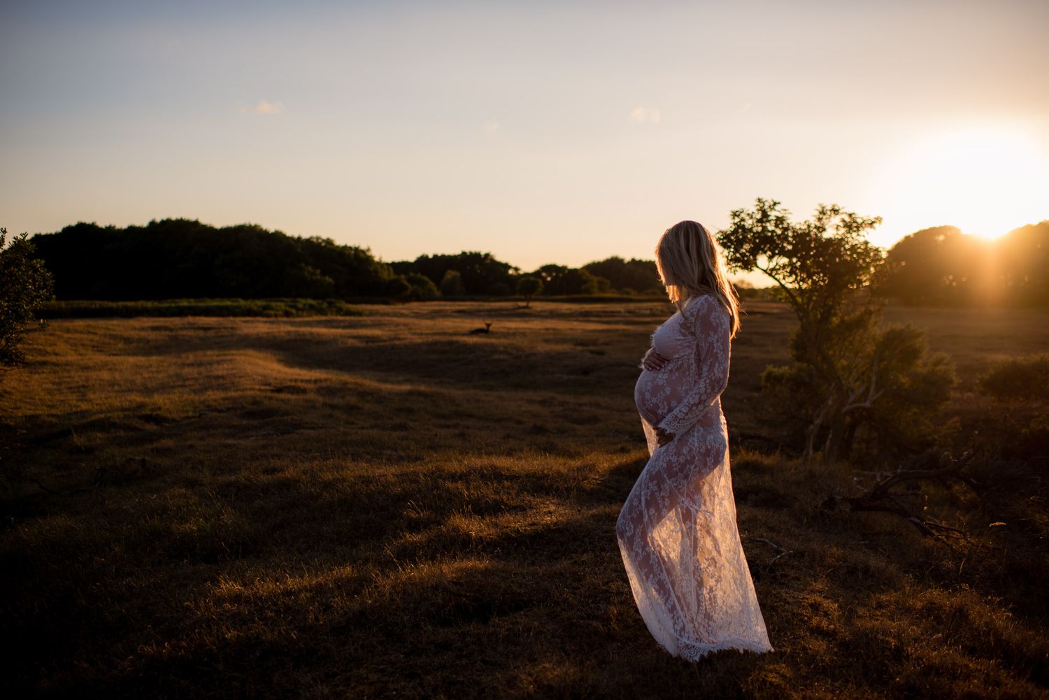 Pregnant woman in white lace dress standing in golden field at sunset.