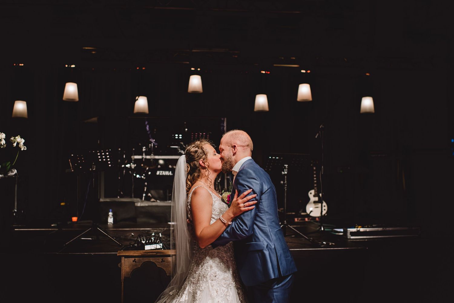 Bride and groom share a romantic first kiss in a dimly lit elegant wedding venue. Door Kim van de Broek Fotografie
