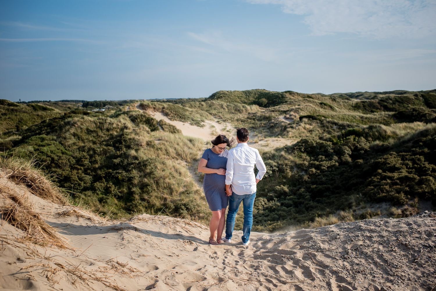Couple walking hand in hand through sandy dunes with green vegetation under a clear blue sky.