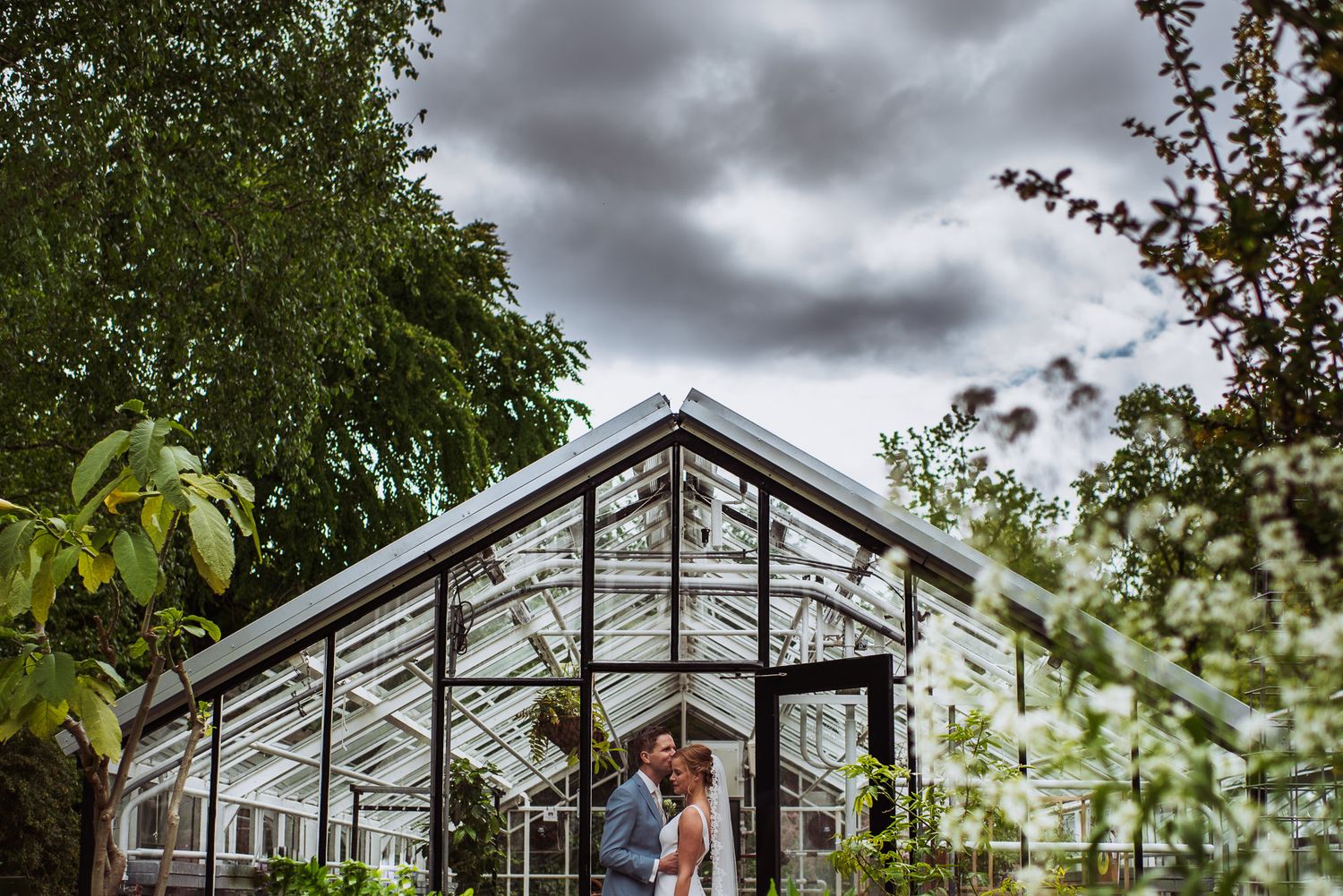 Wedding couple sharing a tender moment in front of a glass greenhouse under cloudy skies.