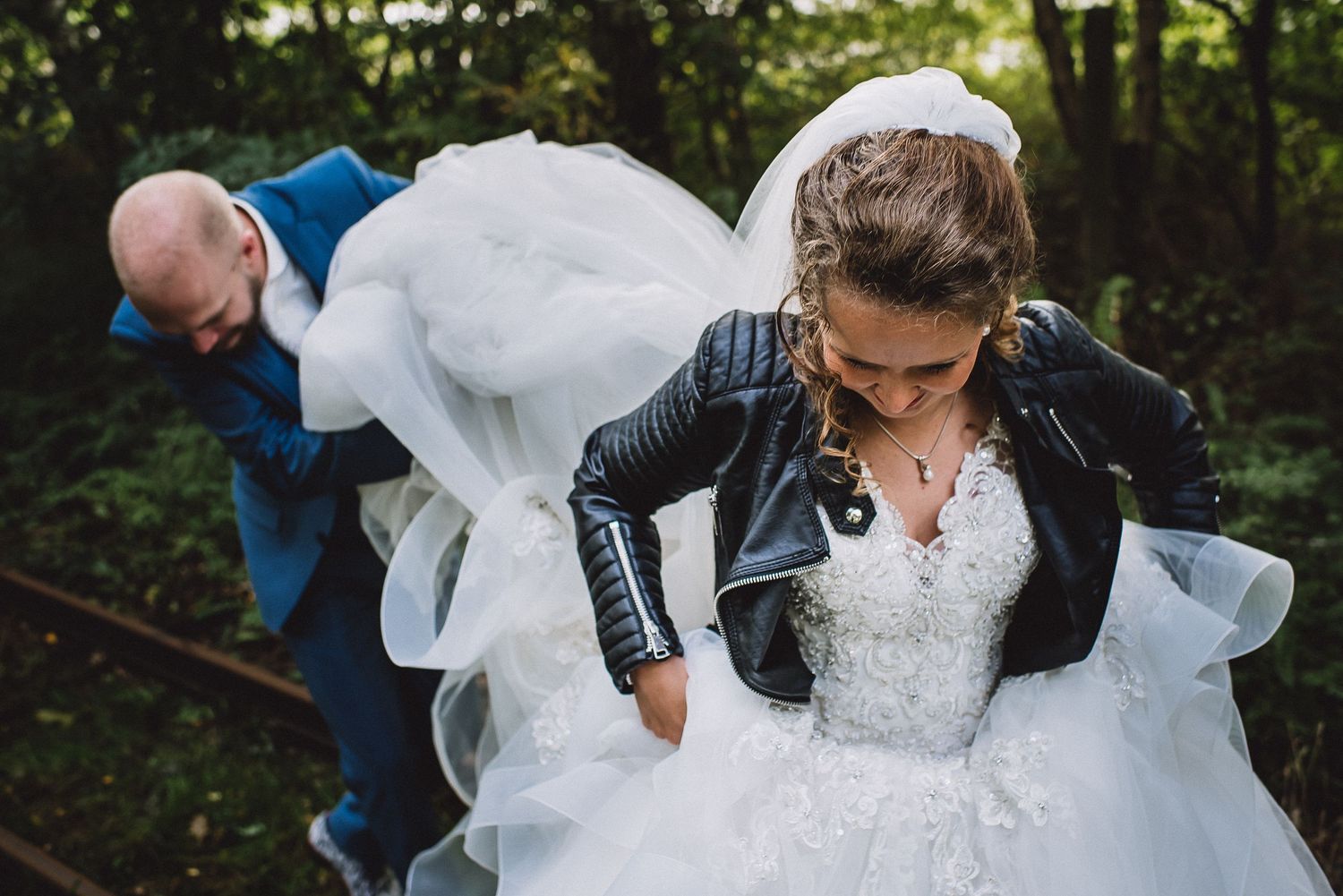 Groom in blue suit lifts bride's white wedding dress train as she adjusts leather jacket in woodland setting.