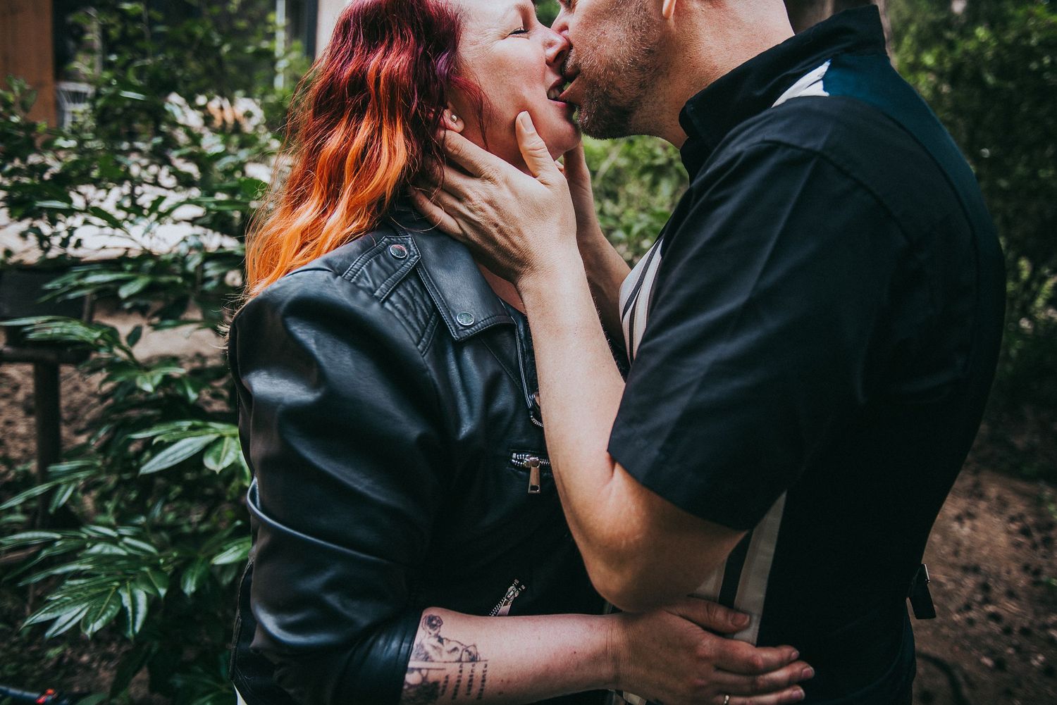 A passionate couple kissing outdoors, the woman with red hair wearing a leather jacket with tattoos.