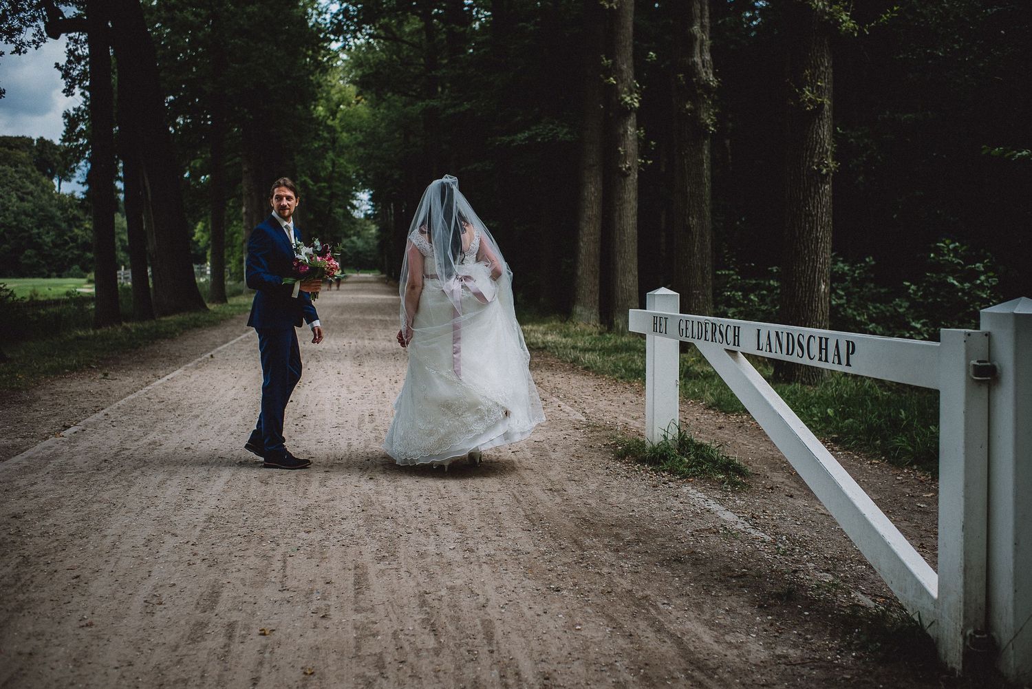 Bride and groom walk hand in hand down a tree-lined path past a white gate on their wedding day.