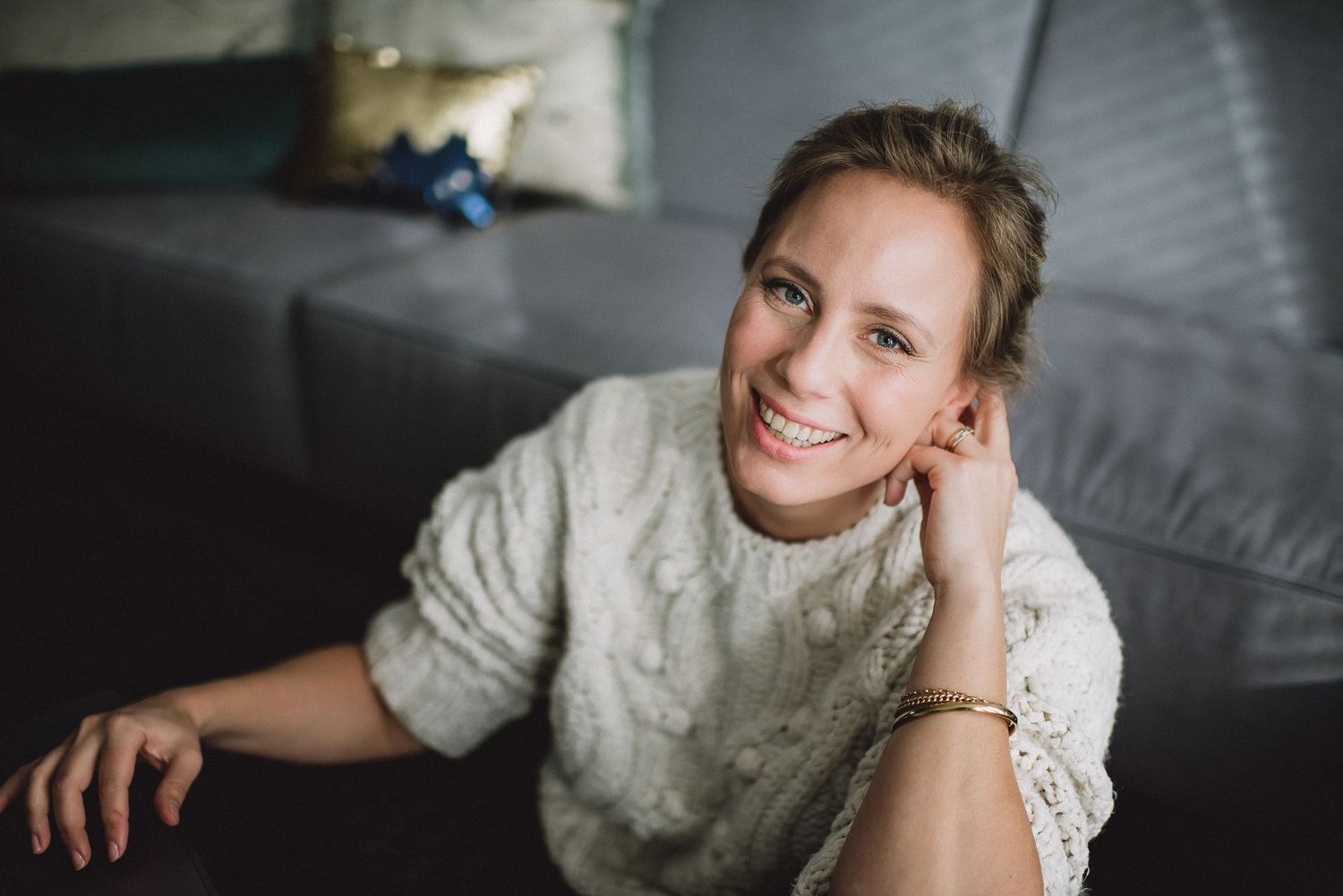 Smiling blonde woman in white knit sweater relaxing on a gray sofa at home.