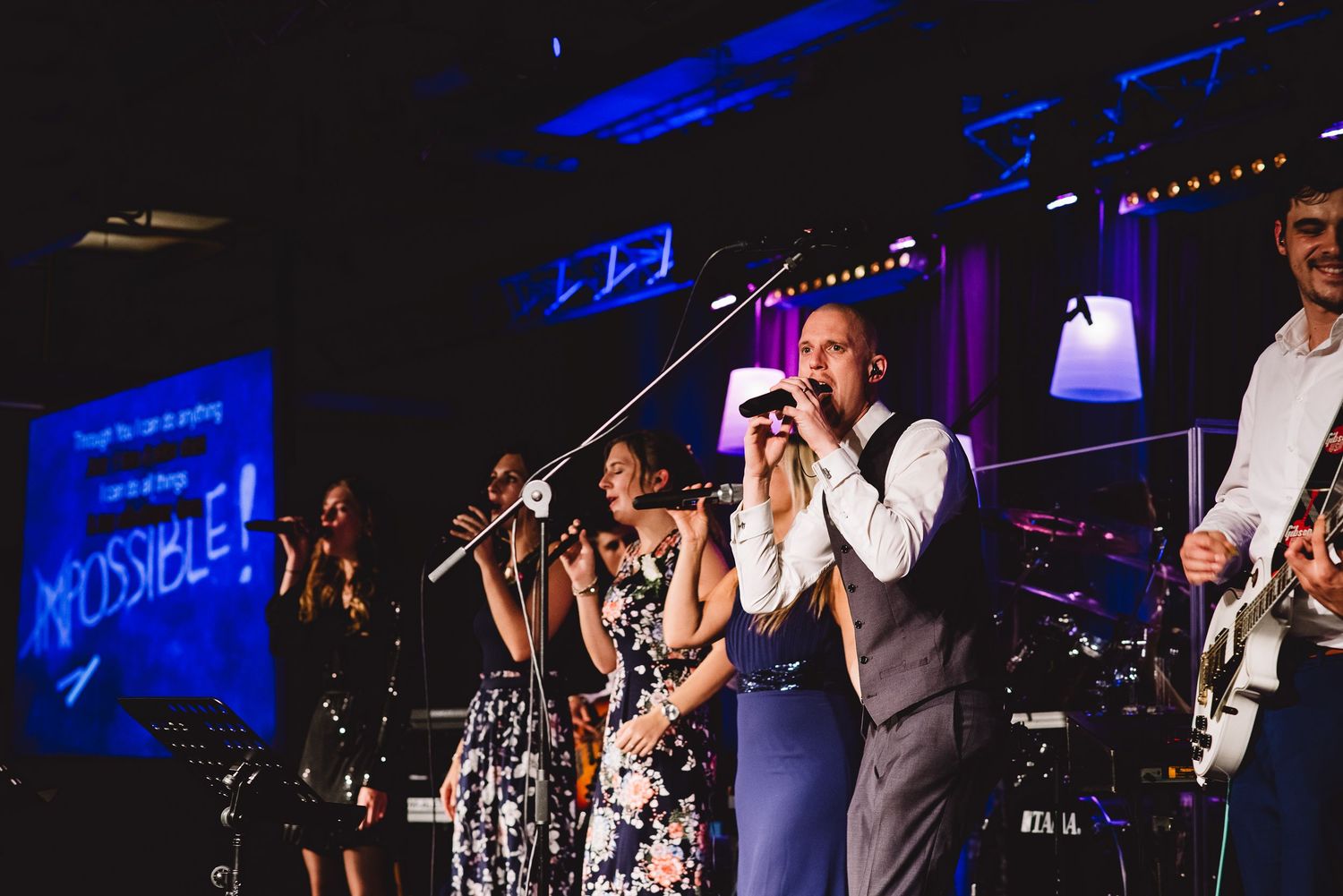 Live band performs on a colorful stage with IMPOSSIBLE displayed on screen at a wedding reception.
