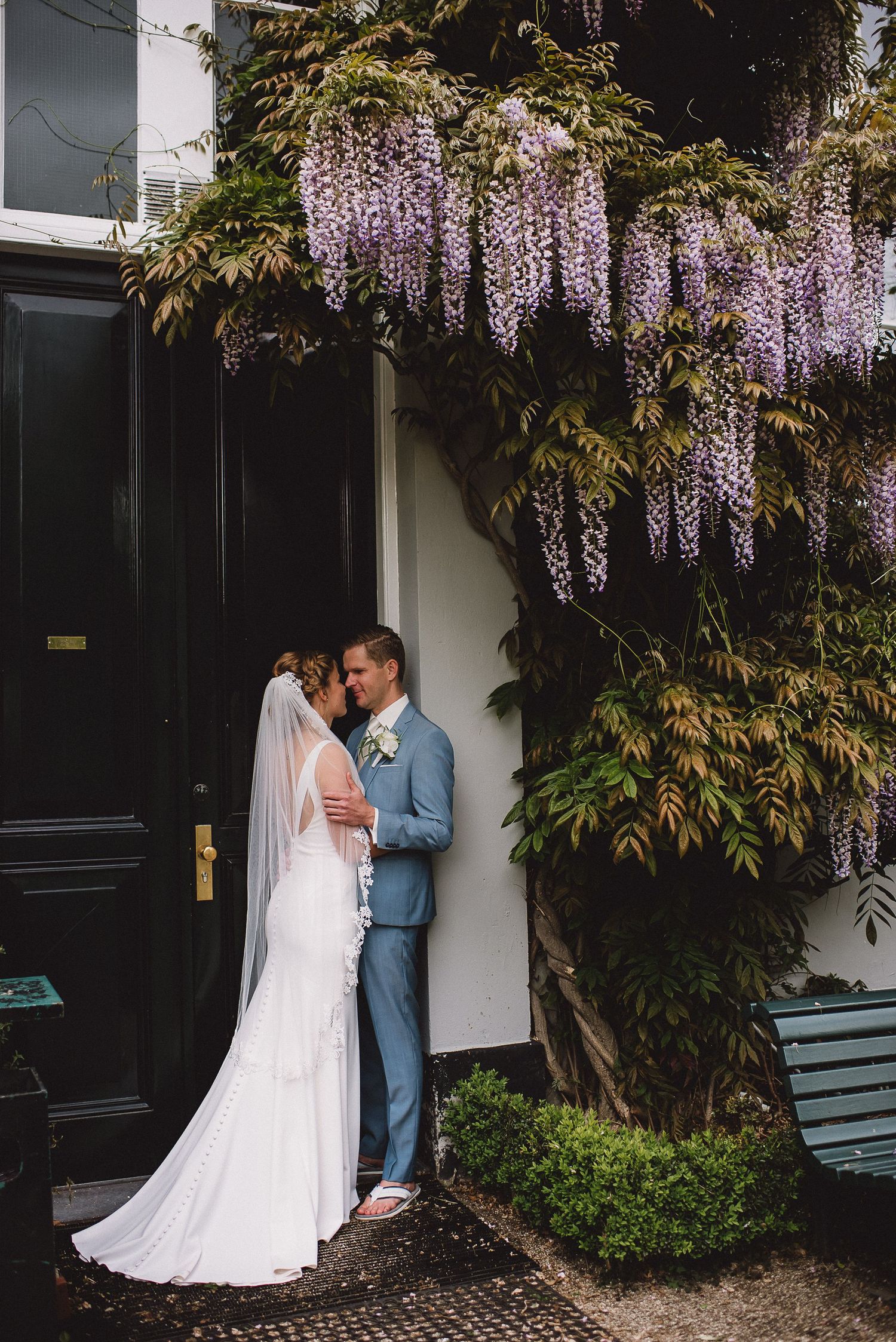 Bride and groom share romantic kiss under cascading purple wisteria flowers on building.Door Kim van de Broek Fotografie