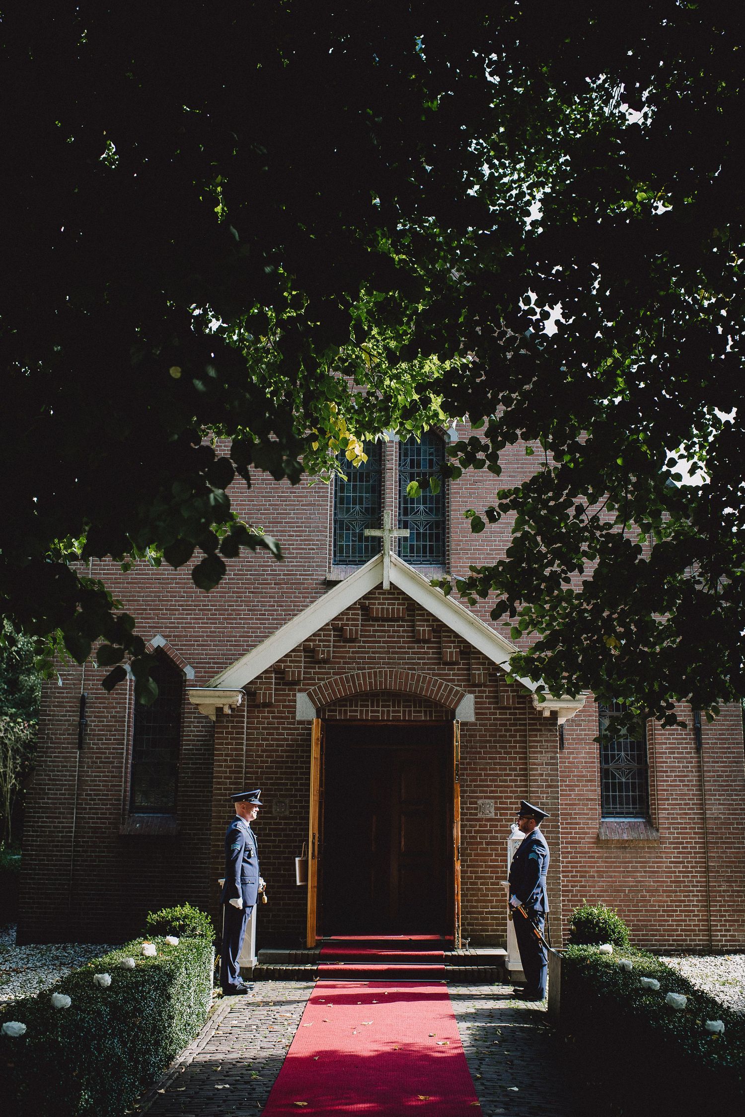Two uniformed guards flanking a red carpet entrance to a brick church with large trees overhead.
