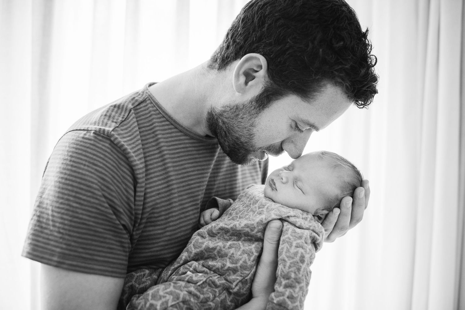 Black and white portrait of a father tenderly touching foreheads with his swaddled newborn baby.
